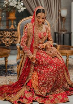 a woman in a red and gold bridal gown sitting on a chair with her hands clasped