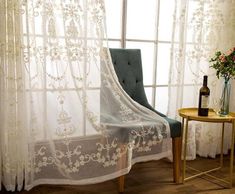 a chair and table in front of a window with sheer curtains on the windowsill