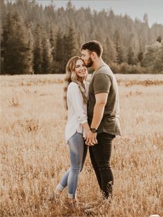 a man and woman standing in a field holding hands while looking at each other with trees in the background