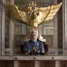 an older man sitting at a desk with two golden clocks in front of him and a chandelier hanging from the ceiling