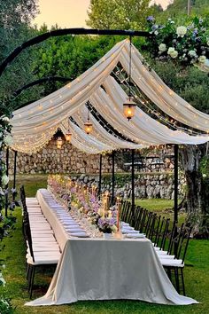 an outdoor dining table set up with white linens and lights on the top, surrounded by greenery