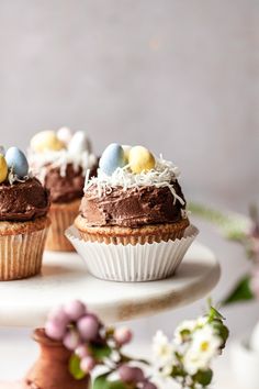 three cupcakes with chocolate frosting and colored eggs on top sitting on a cake plate