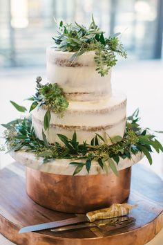 a three tiered wedding cake with greenery on top sits on a wooden platter