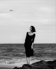 a woman standing on top of a rock next to the ocean in black and white