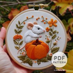 a hand holding up a small embroidered mouse on a pumpkin with leaves and flowers around it