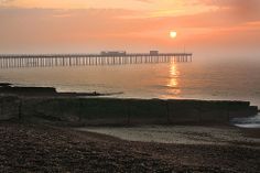 the sun is setting over the ocean with a pier in the distance