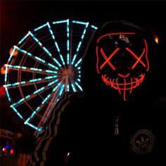 a person wearing a neon mask in front of a ferris wheel with lights on it