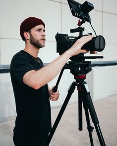 a man standing in front of a camera on a tripod with a red hat