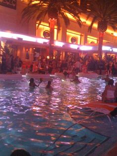 people are in the swimming pool at night with lights on and palm trees behind them