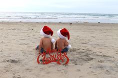 two babies sitting on the beach wearing santa hats with merry christmas signs in front of them