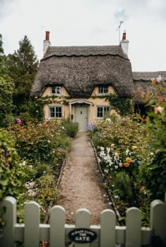 a house with a thatched roof surrounded by flowers and greenery in front of a white picket fence