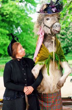 a woman standing next to a large stuffed horse wearing a hat and dress with feathers