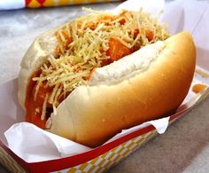 a hot dog with cheese and mustard in a paper container on a counter top next to a napkin