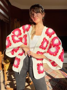 a woman standing in front of a bed wearing a red and white crocheted cardigan