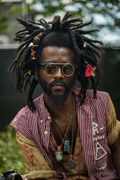 a man with dreadlocks and sunglasses sitting in front of a camera on the ground