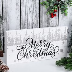 a merry christmas sign sitting on top of a table next to pine cones and evergreens