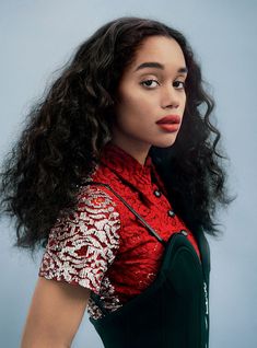 a woman with long curly hair wearing a red shirt and black vest, against a blue background