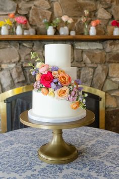 a wedding cake with flowers on it sits on a table in front of a stone wall