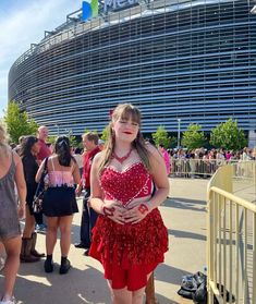 a woman in a red dress standing next to a fence with people walking around her