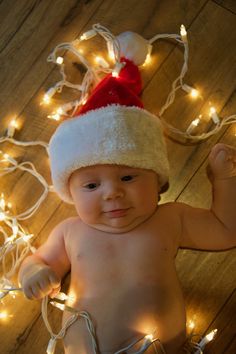 a baby wearing a santa hat on top of a wooden floor with christmas lights around it