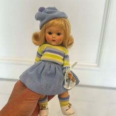 a hand holding a small doll wearing a blue and yellow striped dress with white shoes