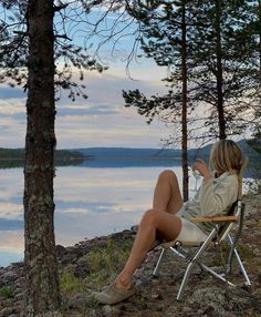 a woman sitting in a chair on the shore of a lake drinking from a cup