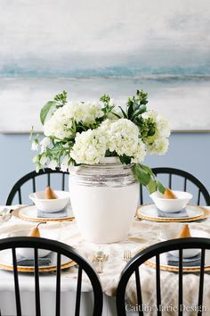 white flowers in a vase on top of a table with black chairs and gold rimmed plates