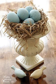 a white vase filled with blue eggs on top of a wooden table next to two small birds