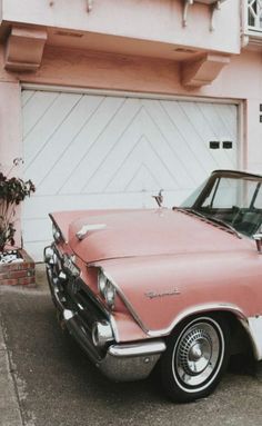 an old pink and white car parked in front of a house with a garage door