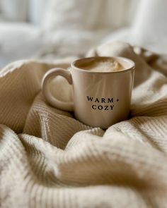 a coffee cup sitting on top of a bed next to a white blanket with the words warm cozy written on it