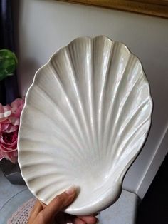 a person holding a white shell shaped dish in front of a flower vase on a table