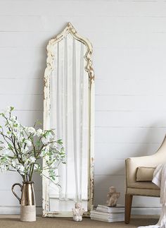 a white chair sitting next to a tall mirror on top of a wooden floor near a vase with flowers