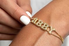 "Introduce our First Retro Gold Pavé Custom Slider Bracelet, and it's absolutely breathtaking.  This bracelet boasts two strands of box chain and letter beads that you can personalize with your name, initials, or favorite phrase.  The Charms are designed in a retro bubble letter font and feature intricate pavé detail, which gives it a luxurious touch.  Moreover, the pull-tie closure makes it simple to adjust to your preference.  You can wear this bracelet every day, and it'll undoubtedly become a staple in your jewelry collection.  Make sure to explore the Letter Key to see all the options for customization! * Material: High-quality and Anti Tarnish 14K Gold Plated - Brass; AAA Grade Cubic Zirconias. * Finish: Up to 5.75\" (14.5 cm) in diameter, adjustable 14K Gold Plated Box Chain.  HOW T Name Bracelet Gold, Bubble Letter Fonts, Bracelet Name, Gold Bubbles, Bubble Letter, Name Initials, Dope Jewelry, Gold Bracelet For Women, Funky Jewelry