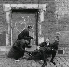 black and white photograph of three men sitting on chairs in front of a brick building