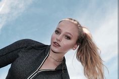 a woman with long hair standing in front of a blue sky wearing a black jacket