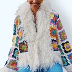 a woman sitting on top of a chair wearing a white jacket and colorful crocheted sweater