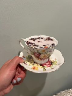 a person holding a tea cup and saucer with flowers on it, in front of a gray wall