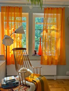 a rocking chair in front of two windows with orange drapes on the window sill