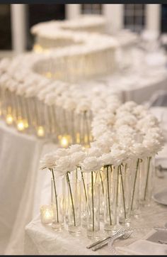 white flowers in vases are lined up on a table with silverware and candles