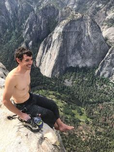 a shirtless man sitting on top of a mountain next to a valley filled with trees