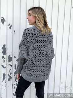 a woman standing in front of a white wall wearing a gray crocheted sweater