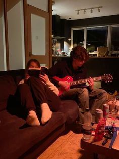 two people sitting on a couch playing guitar in the living room, one person is holding a cell phone