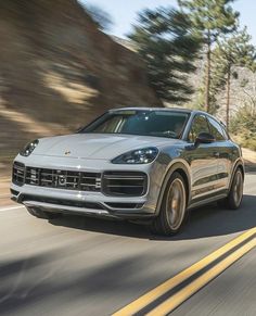 a silver porsche cayen is driving down the road with trees in the back ground