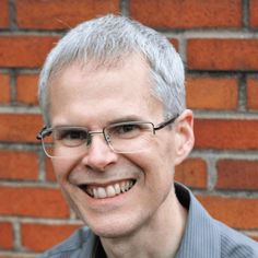 a man with glasses smiling in front of a brick wall