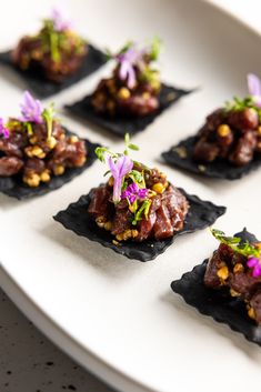 small pieces of food are arranged on black slates with purple flower garnishes