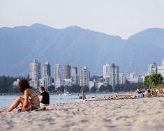 people are sitting on the beach in front of some mountains and water with buildings behind them