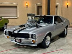 a silver muscle car parked in a garage next to a green mustang truck with an engine on it's hood