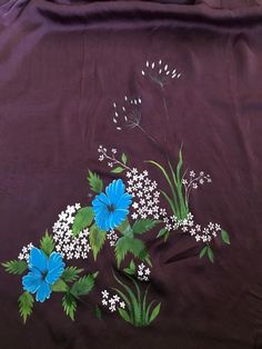 an embroidered cloth with blue flowers and white daisies on brown fabric, as well as green leaves