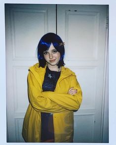 a woman with blue hair standing in front of a white door wearing a yellow jacket