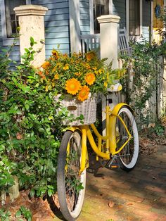 a yellow bicycle with flowers in the basket parked on a brick walkway next to a blue house